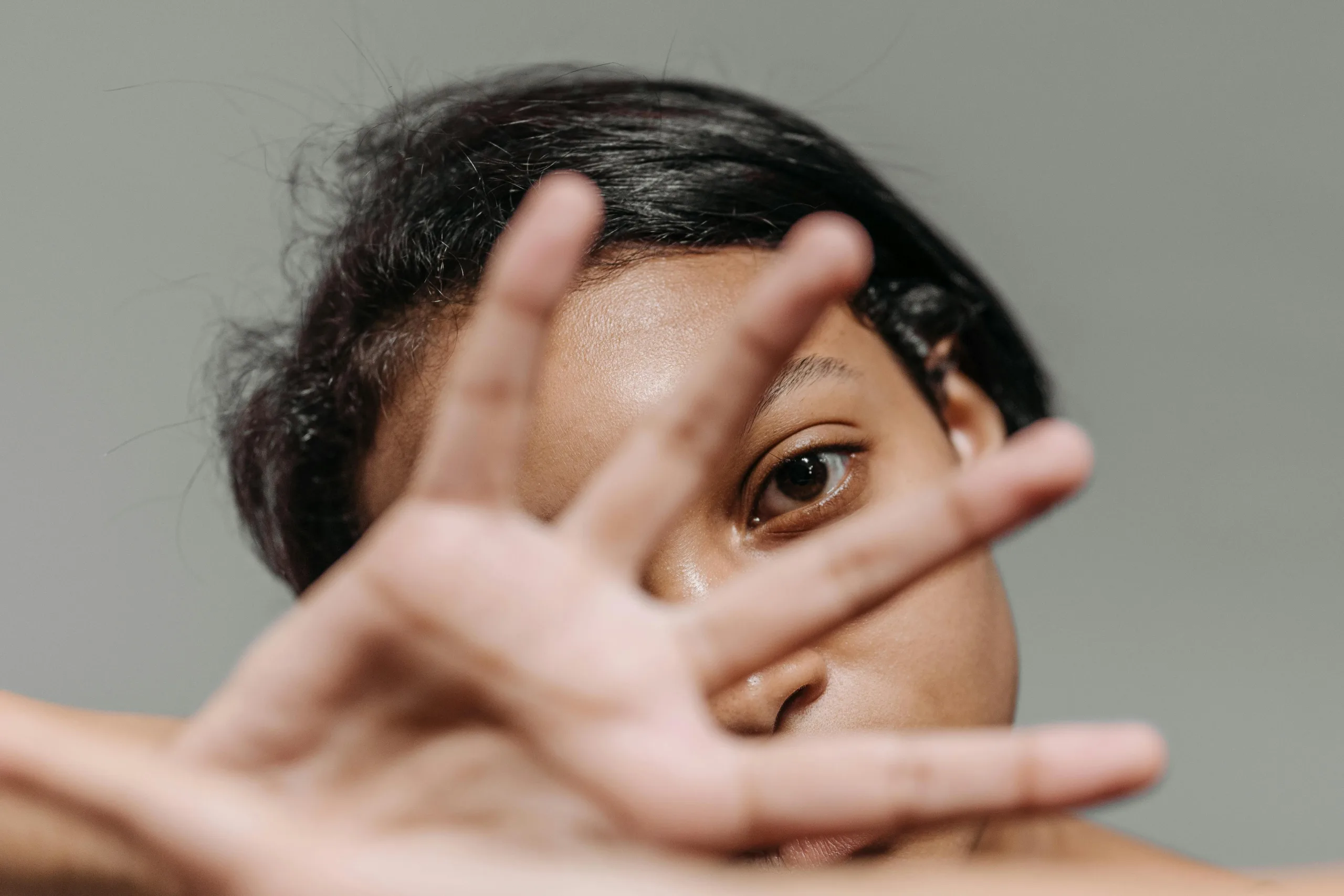 Intimate portrait of a woman partially covering her face with her hand against a gray background.